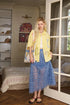 Woman in a yellow shirt and blue skirt standing in a room with a bed and window.